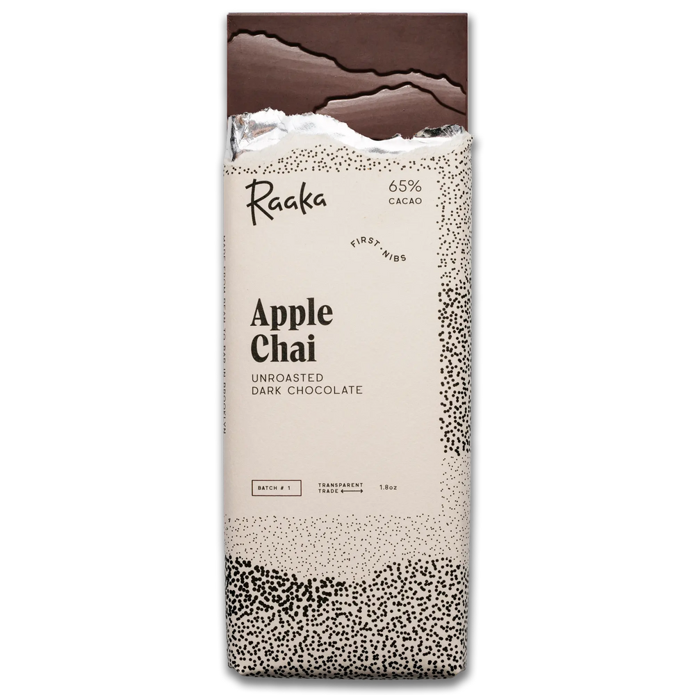 A vertical pack of Raaka Apple Chai unroasted dark chocolate bars, featuring a cream-colored wrapper with black text and speckled patterns, and a dark chocolate bar broken at the top, on a plain white background.
