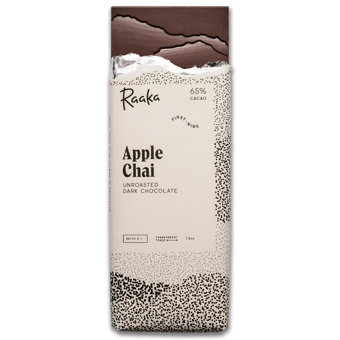 A vertical pack of Raaka Apple Chai unroasted dark chocolate bars, featuring a cream-colored wrapper with black text and speckled patterns, and a dark chocolate bar broken at the top, on a plain white background.