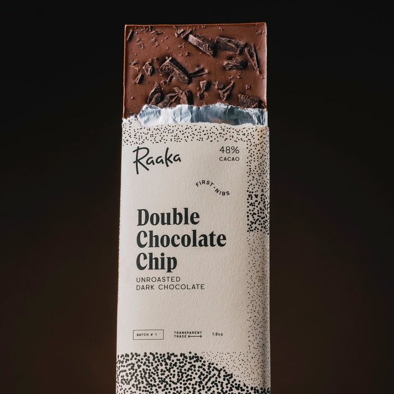 A single upright bar of Raaka Double Chocolate Chip in its paper wrapper, shown against a dark gradient background. The wrapper is cream-colored with black speckled patterns and text, and the top section of the chocolate bar is visible, showing a rich dark brown chocolate with chocolate shavings.