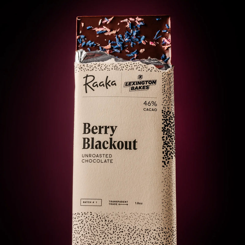 A packaged chocolate bar from Raaka and Lexington Bakes, called Berry Blackout. It's a 46% cacao unroasted chocolate. The wrapper is cream-colored with black speckled patterns and text, and the top of the transparent section shows dark chocolate with blue and pink coconut sprinkle pieces.