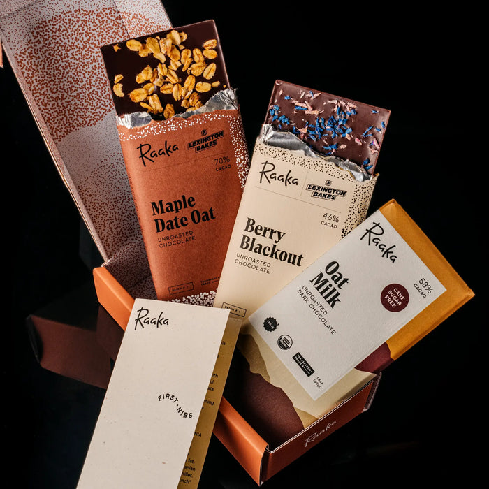 A box of Raaka chocolate bars arranged in an open orange box against a black background. Visible bars include Maple Date Oat, Berry Blackout, and Oat Milk.