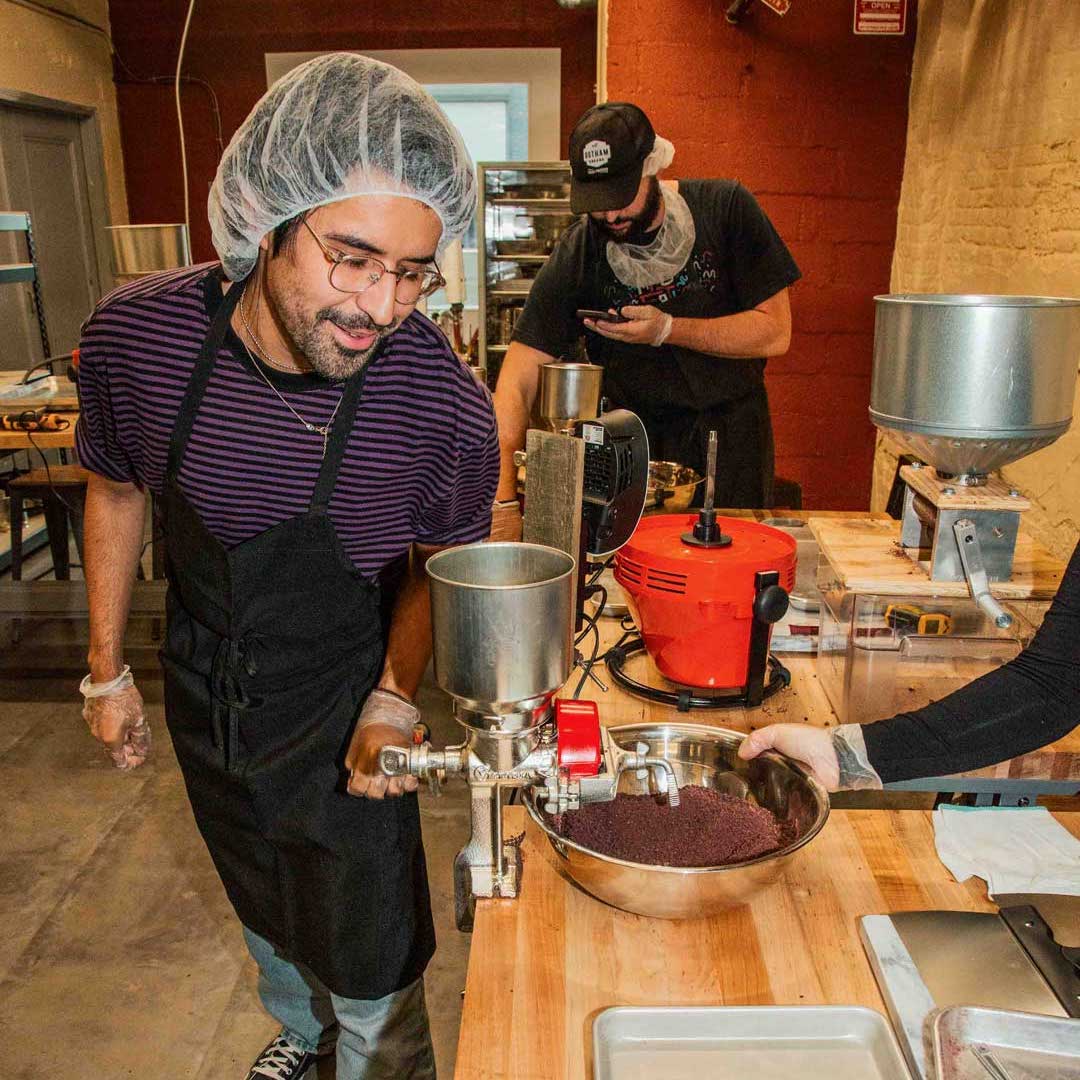 A man in a hairnet using a hand grinder to grind cacao beans into a bowl. 